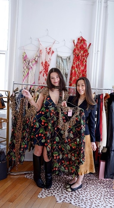 Two VYA sellers at a pop-up, one holding up a cherry-print vintage dress in front of a rack of floral dresses